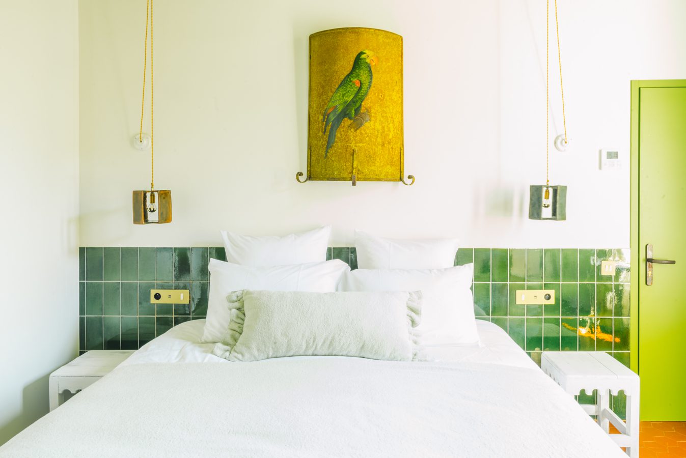 The green suite at La Relève hotel, showcasing a vintage-inspired bed with green-tiled accents, a unique parrot wall light, and gold pendant lamps.