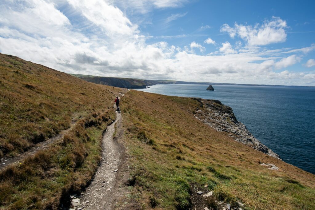 A hiker walking along the South West Coast Path, a scenic cliffside trail managed by the National Trust, with views of the rugged coastline, the blue sea, and a distant rock formation under a partly cloudy sky.