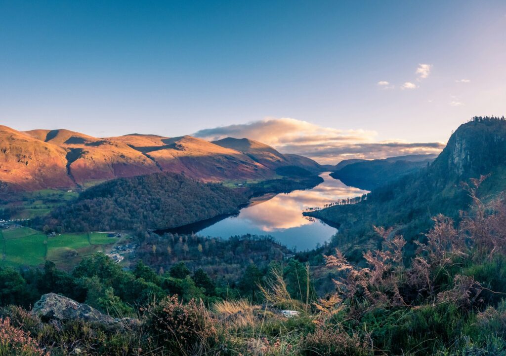 A breathtaking view of the Peak District, featuring rolling hills bathed in golden sunlight, a reflective lake nestled between valleys, and a clear blue sky with scattered clouds.