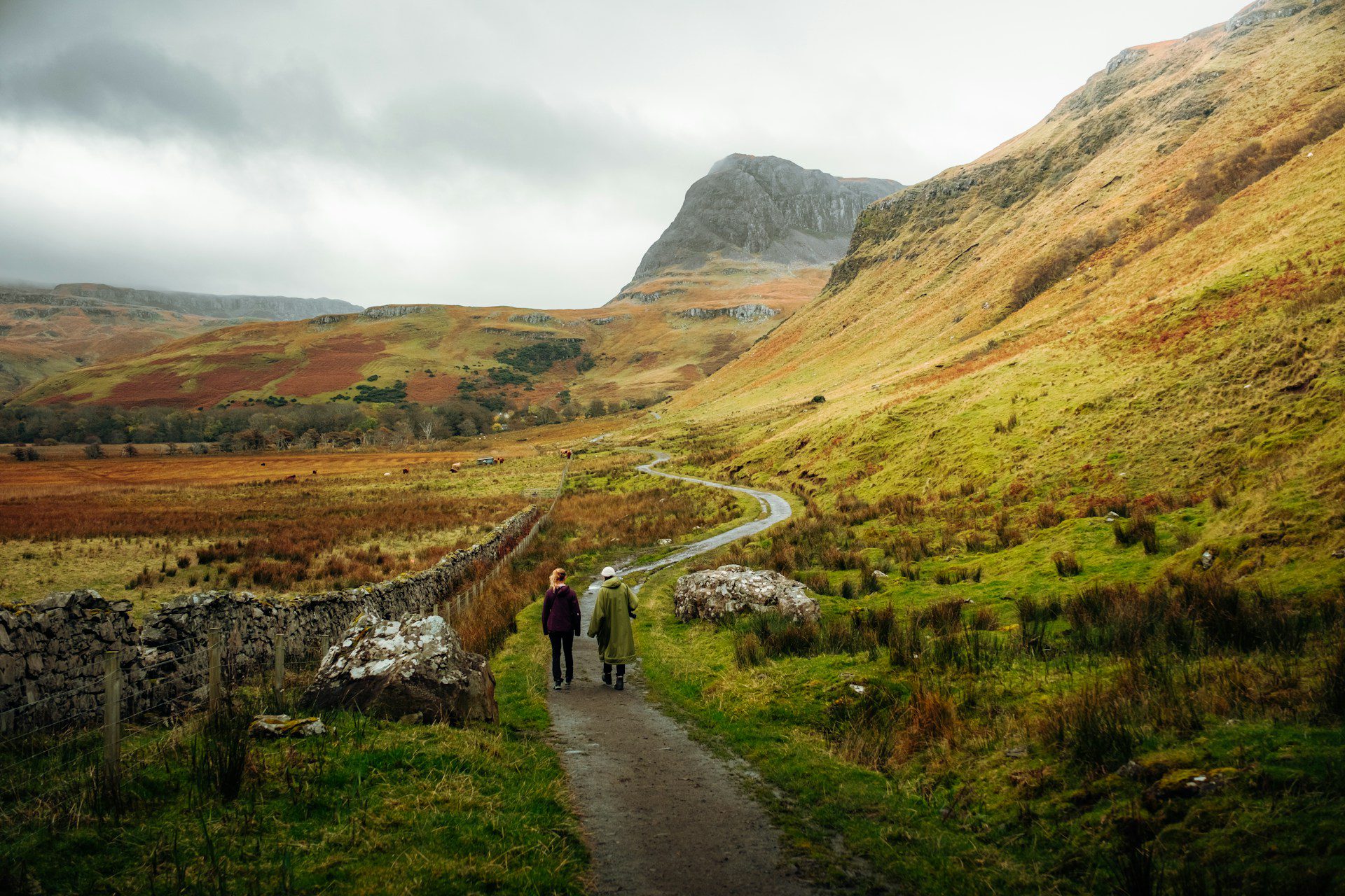 Two hikers walking along a winding trail in the Isle of Skye, Scotland, surrounded by rolling green hills, rugged mountains, and a dramatic cloudy sky.