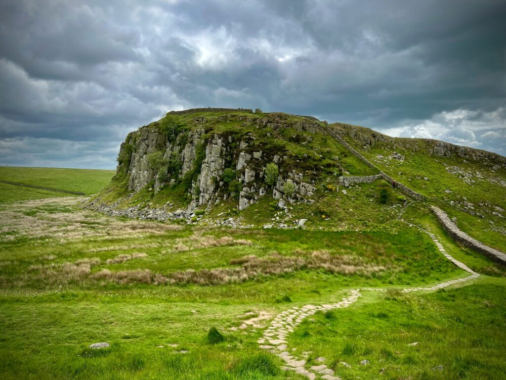 A scenic view of Hadrian’s Wall in Northumberland, stretching up a craggy hillside with ancient stone fortifications surrounded by lush green fields under a dramatic cloudy sky.