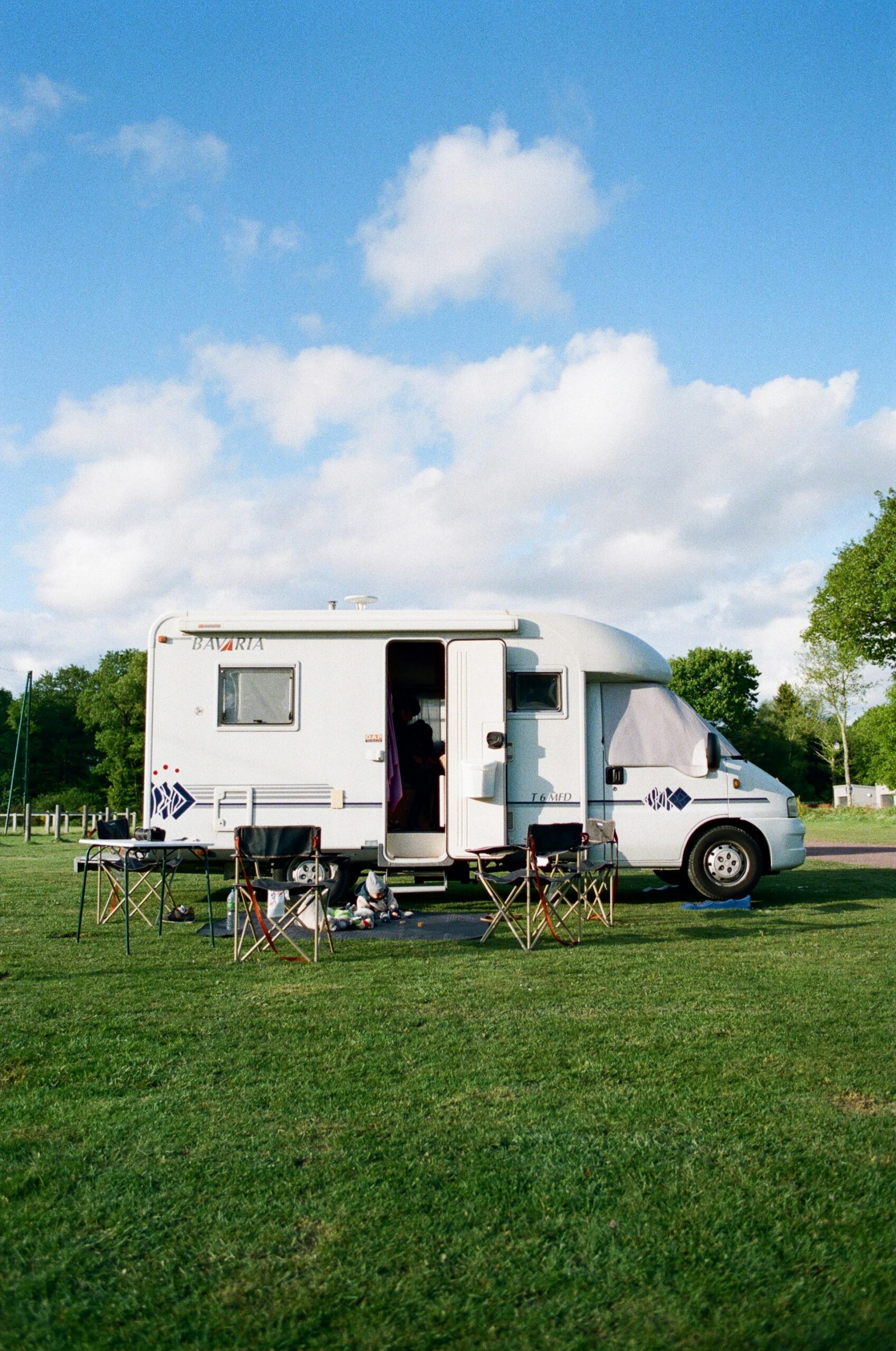 A white campervan with an open door is parked on lush green grass under a bright blue sky with scattered clouds. Several foldable camping chairs and tables are arranged outside, creating a relaxed outdoor setup.