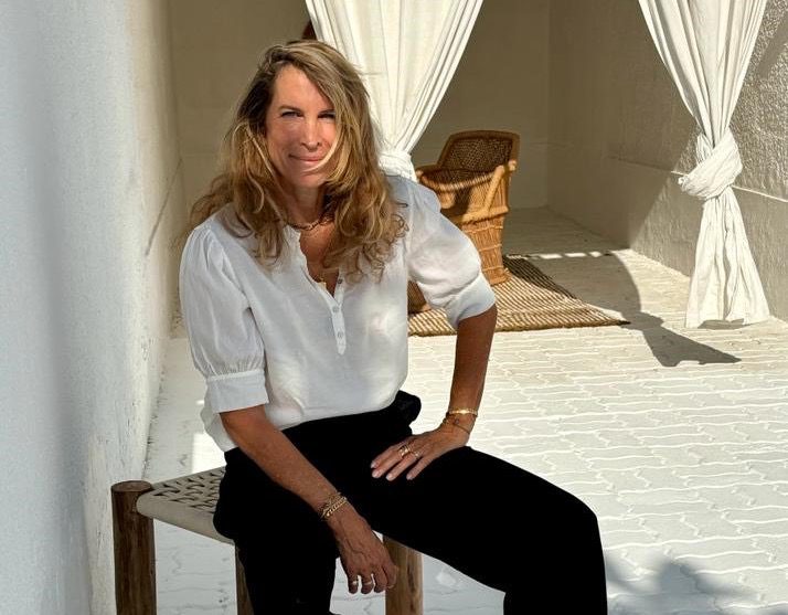 Mathilde Rottembourg seated on a wooden stool in a sunlit, white-washed patio, wearing a white blouse and black trousers.
