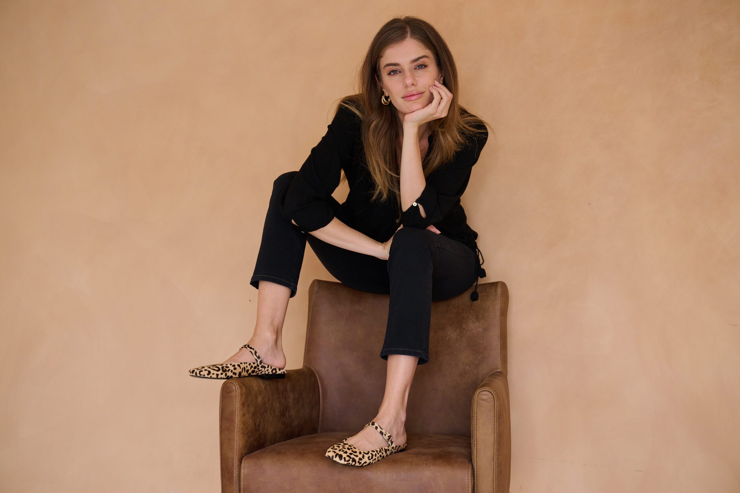 Woman in all-black sitting on a leather chair, showcasing leopard-print ballerina mules by With My Sands.