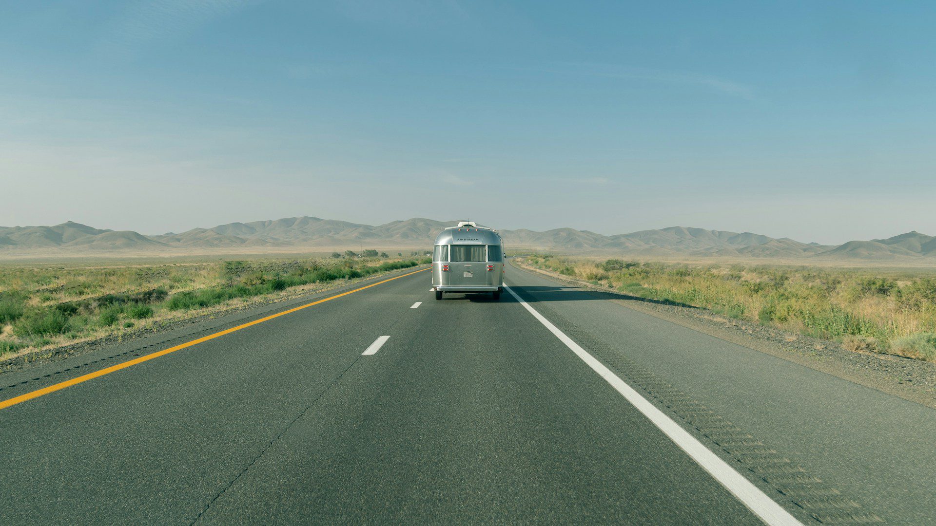 Silver Airstream trailer driving down an empty desert highway, with distant mountains under a clear blue sky.