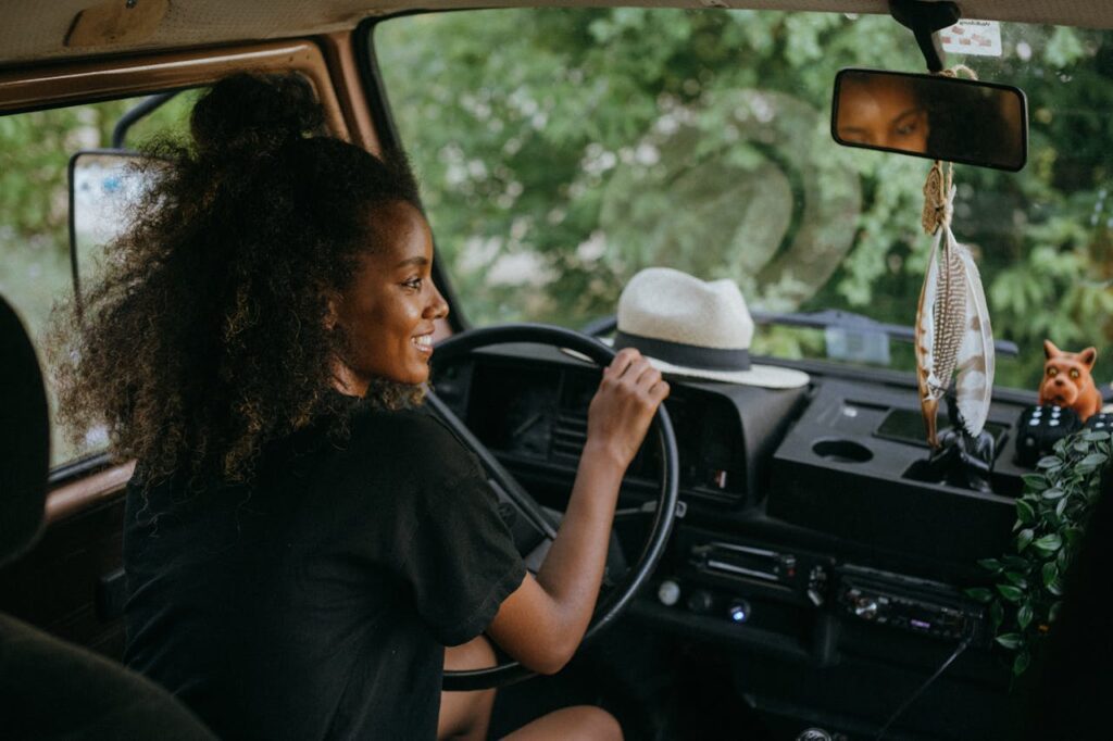 A woman smiling while driving a vintage van, with a hat on the dashboard and greenery outside the window.