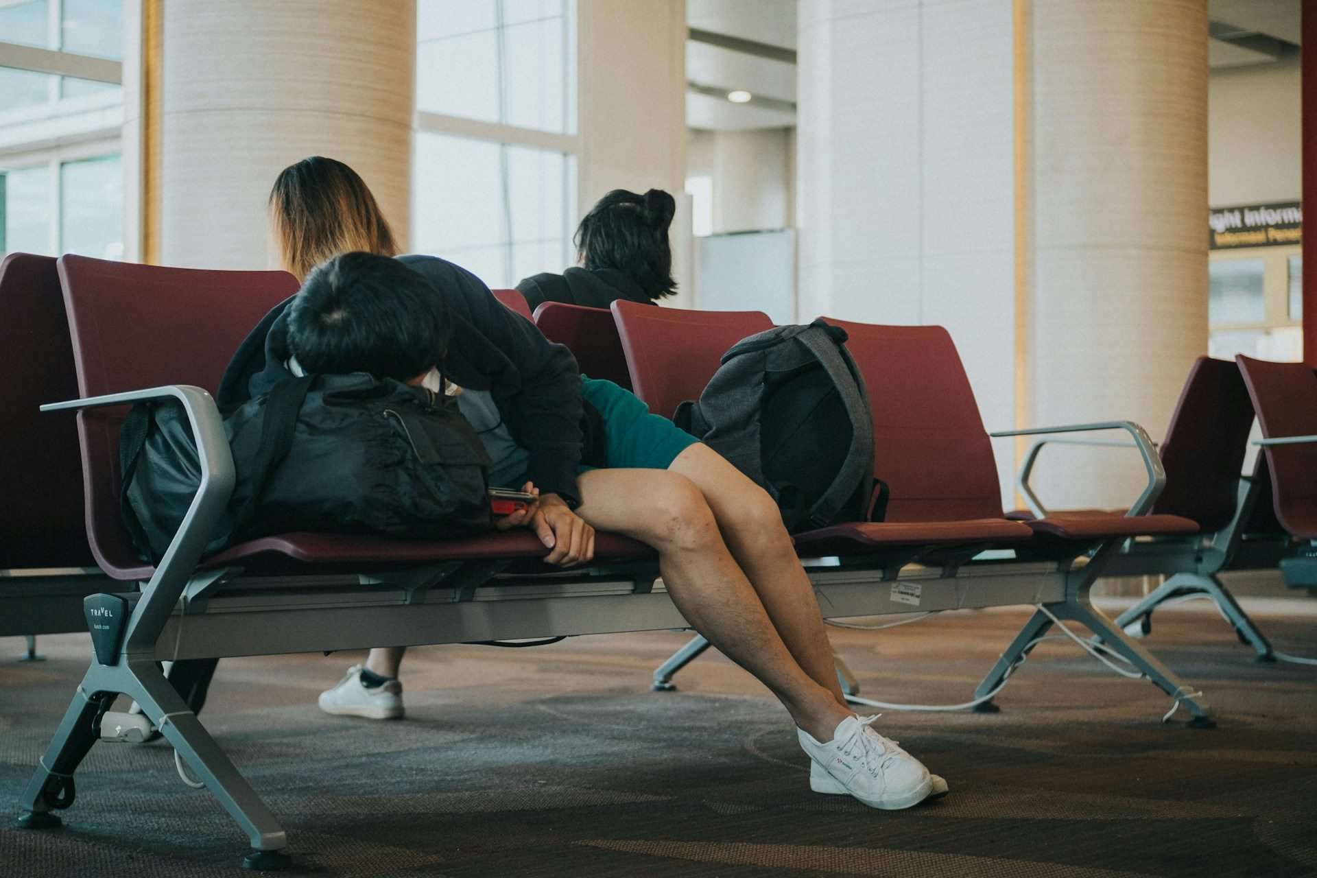 A traveller asleep on airport seating, backpack used as a pillow, highlighting exhaustion from flight delays.