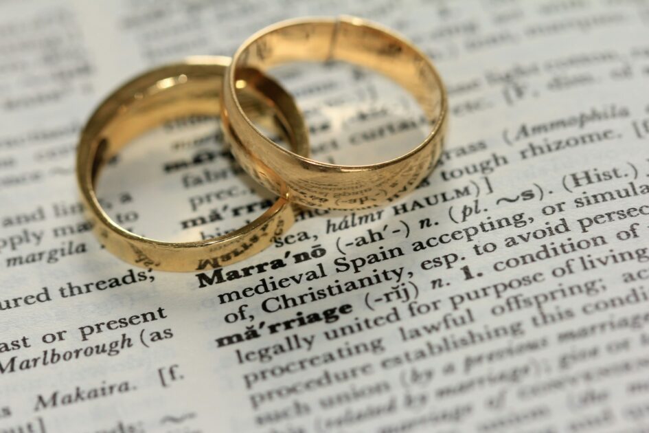 Close-up of two gold wedding bands resting on a dictionary page open to the word “marriage,” symbolising love, union, and lifelong commitment.